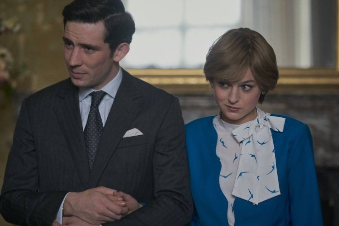 A imagem mostra uma cena da série The Crown, na qual o príncipe Charles, um homem branco com os cabelos curtos e pretos, usando um terno preto, com camisa branca e gravata cinza, além de um lenço branco no bolso, está de braços dados com Diana, uma mulher branca, loira e com cabelos curtos, usando um blazer azul e uma camisa branca, com detalhes em azul e um laço na gola, por baixo do blazer.